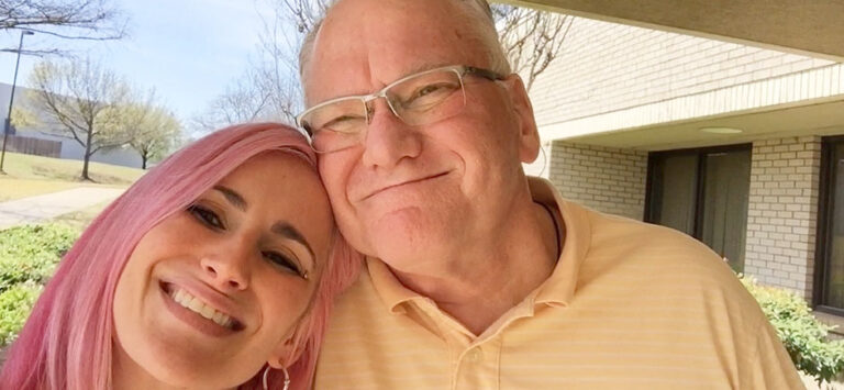 A smiling woman with pink hair poses closely with an older man, both enjoying a sunny day outdoors.