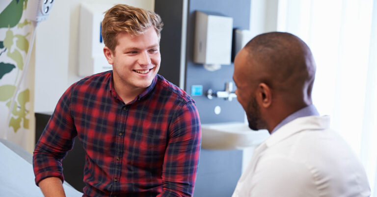 A young man in a plaid shirt smiles while talking to a healthcare professional in a medical setting.