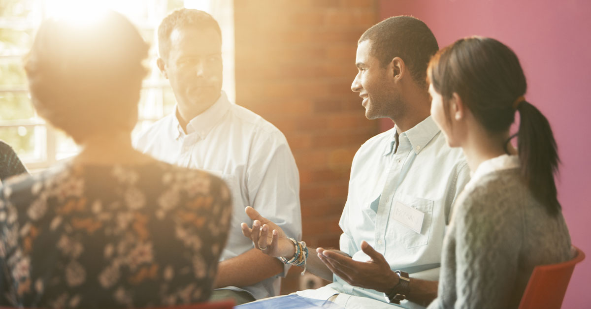 A group of people engaged in conversation, with a focus on two individuals sharing ideas in a warm, inviting setting.