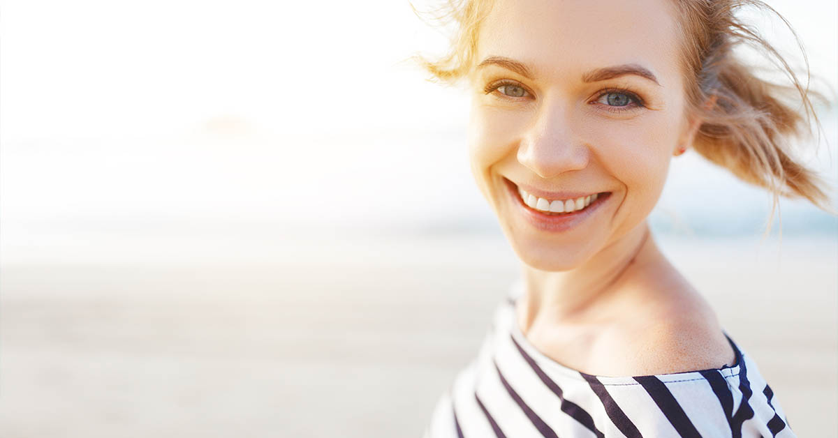 A smiling woman with wavy hair stands on a beach, wearing a striped top, with a bright, sunny background.
