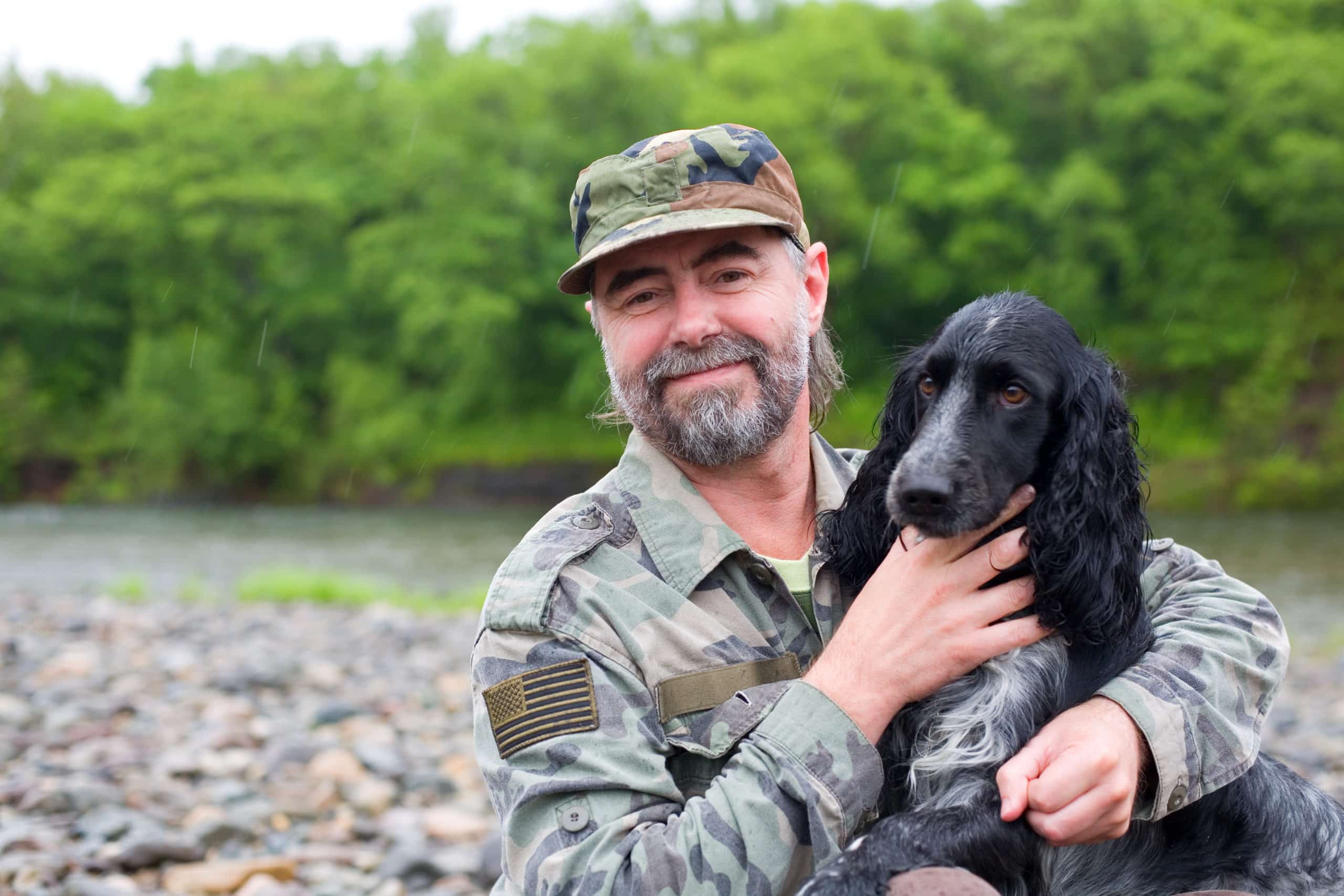A man in military camouflage holds a black dog by a river, surrounded by lush greenery.