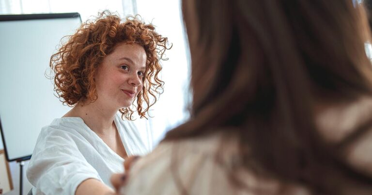 A woman with curly hair smiles while reaching out for a handshake in a bright, modern setting.