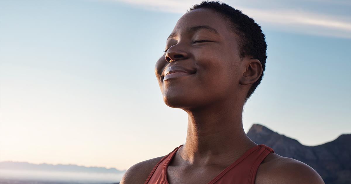 A person with short hair smiles peacefully against a scenic outdoor backdrop, enjoying the fresh air.