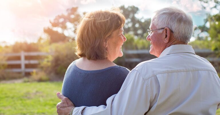 A couple stands together, smiling at each other, with a sunny outdoor background and a fence in the distance.