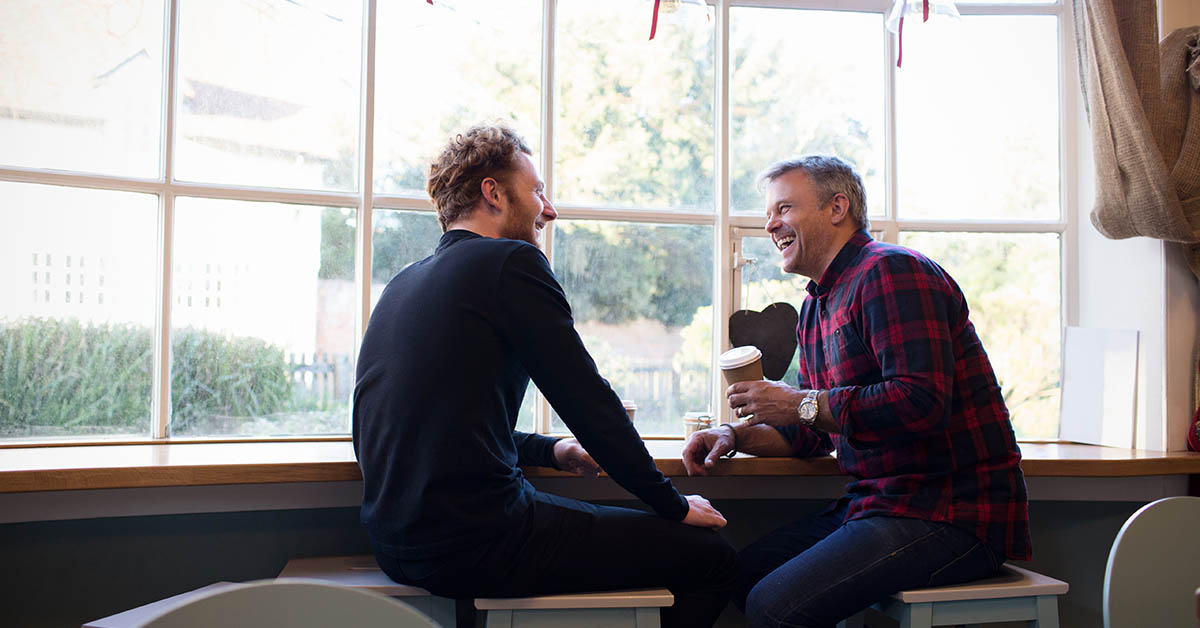 Two men are sitting by a window, laughing and enjoying coffee together in a cozy setting.