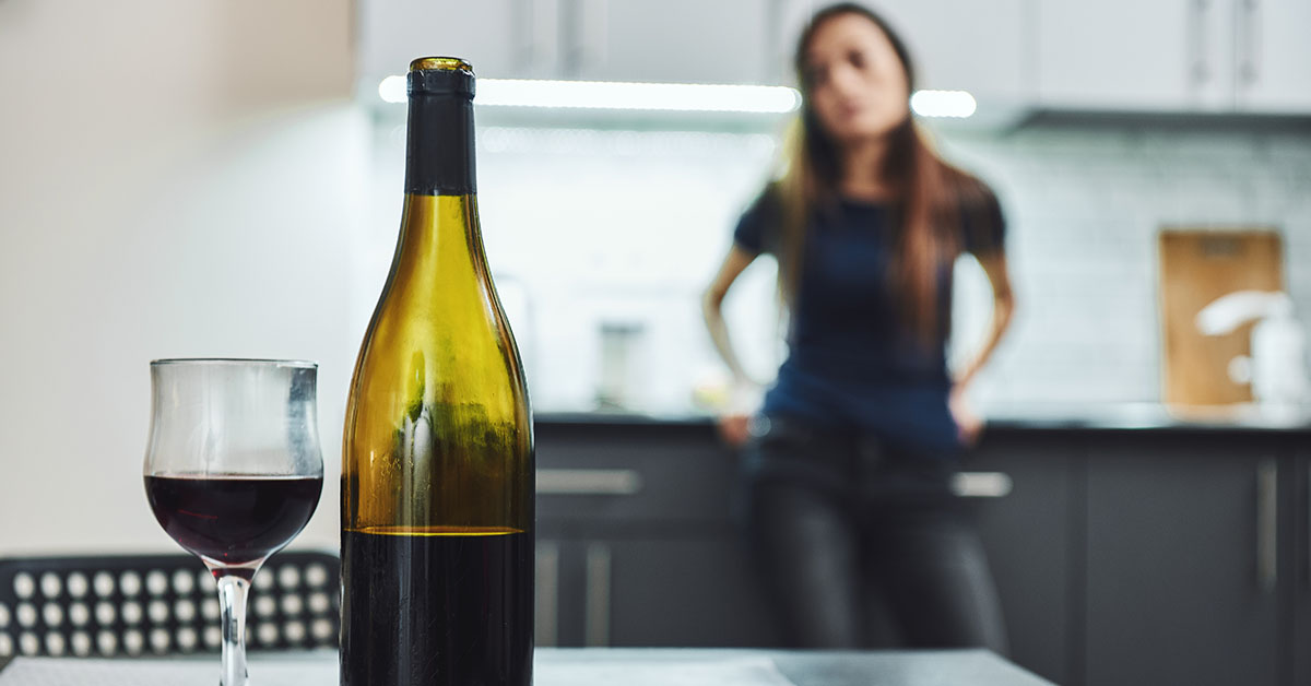 A wine bottle and glass are in focus on a table, with a blurred figure of a person standing in a kitchen background.