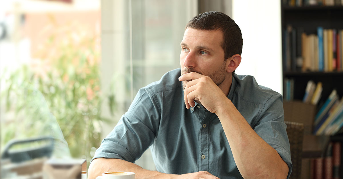 A man in a blue shirt sits pensively at a table, resting his chin on his hand, with a coffee cup nearby.