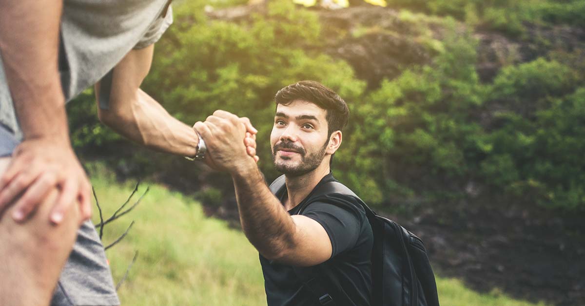 Two men are helping each other on a hike, with one reaching out to assist the other. Lush greenery surrounds them.