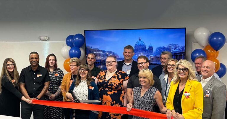 A group of people gathers for a ribbon-cutting ceremony, smiling and holding a large red ribbon. Balloons decorate the background.