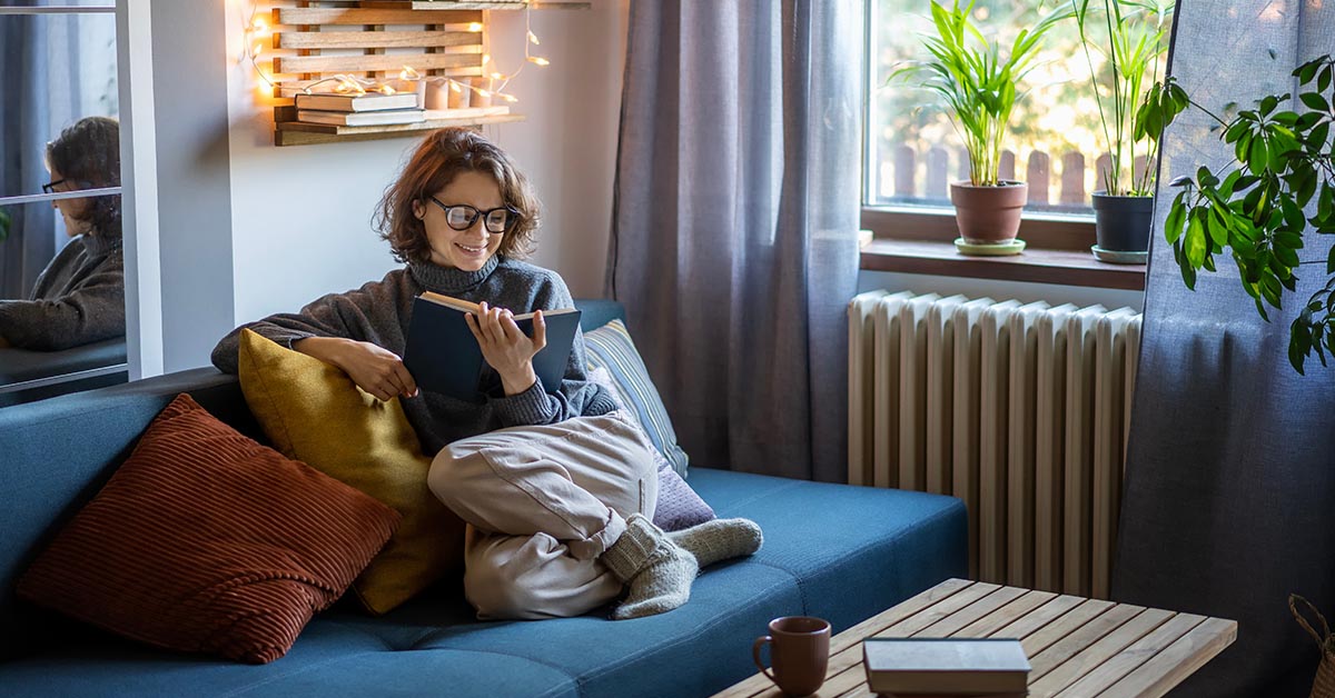A cozy living room scene with a woman reading on a blue couch, surrounded by plants and warm lighting.