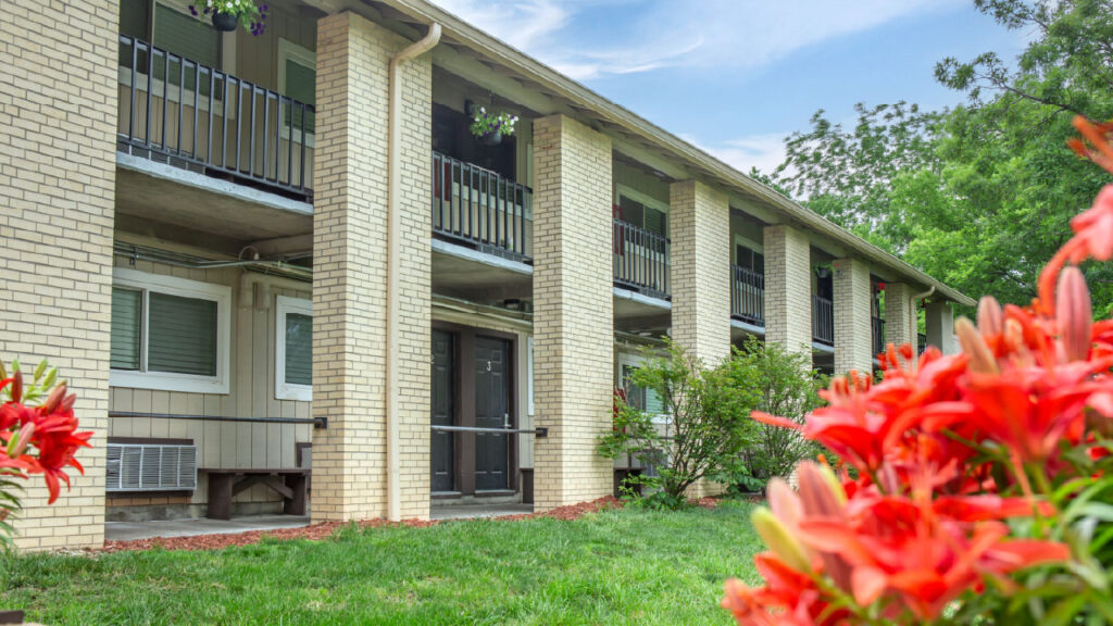 A multi-story apartment building with balconies, surrounded by green grass and vibrant red flowers in the foreground.
