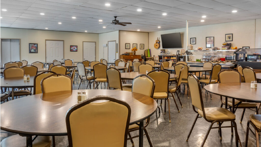 A spacious dining area with round tables and beige chairs, featuring a serving counter and wall decorations.