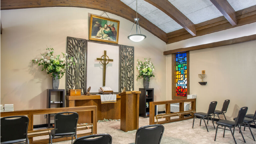A serene chapel interior featuring a wooden altar, a cross, floral arrangements, and a stained glass window. Chairs are arranged for seating.