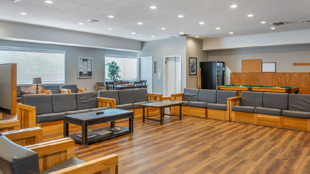 A spacious lounge area with wooden furniture, gray sofas, a coffee table, and a pool table in the background. Brightly lit with large windows.