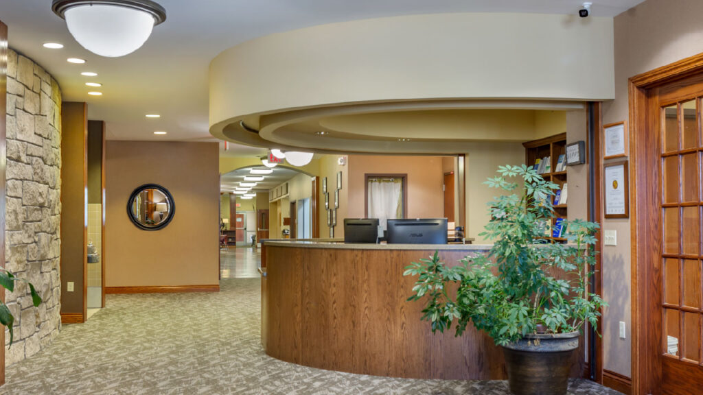 A modern reception area featuring a curved wooden desk, stone wall, and decorative plants. Bright lighting and a welcoming atmosphere.