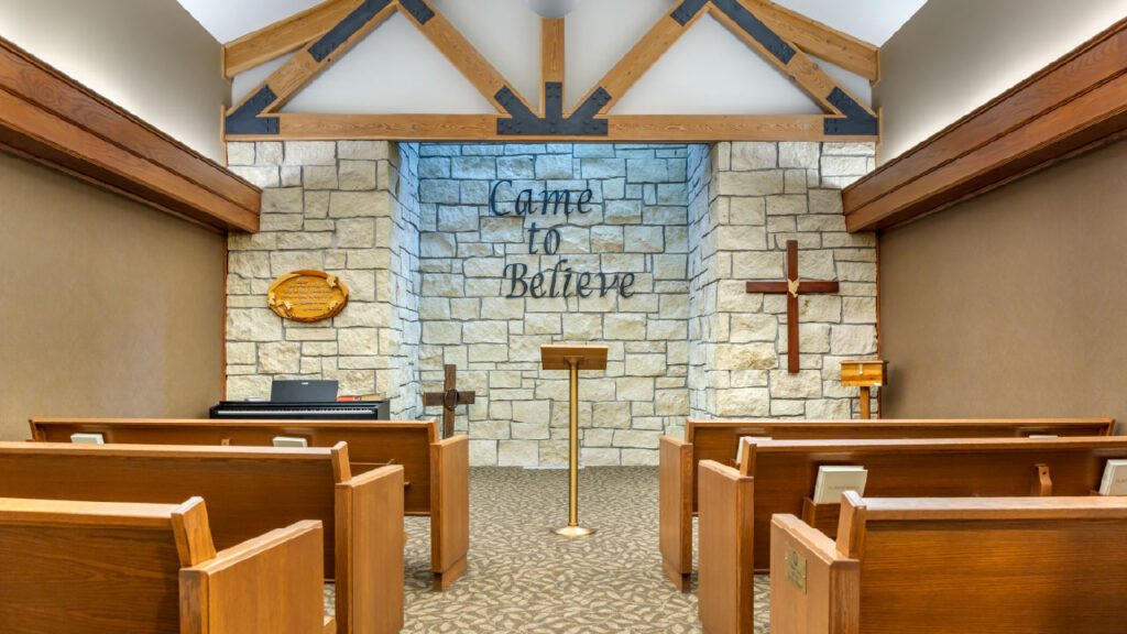A serene chapel interior with wooden pews, a stone wall, a podium, and religious symbols, featuring the phrase "Came to Believe."