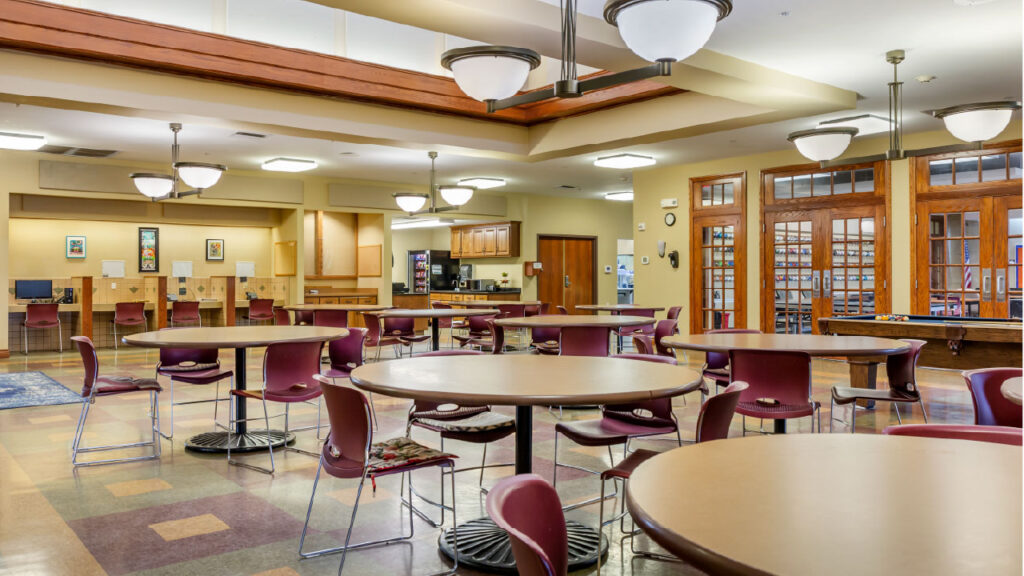A spacious communal area with round tables and chairs, featuring a kitchenette and computer stations in a warm, inviting setting.