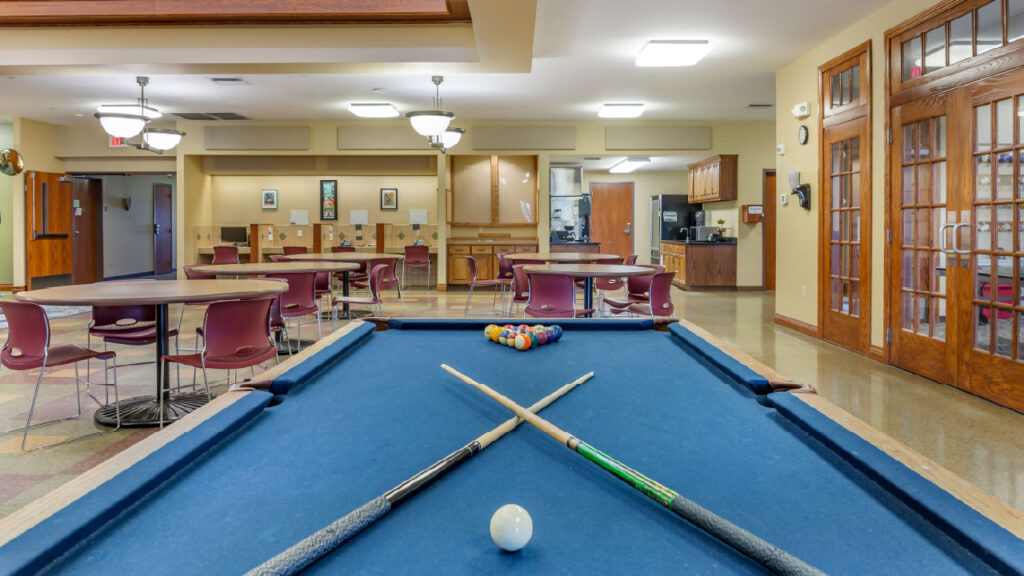 A pool table with cues and balls in a spacious recreational room featuring tables and chairs, with a kitchenette in the background.