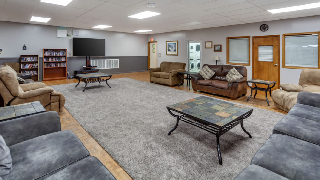 A spacious lounge area with multiple sofas, a coffee table, a TV, and a bookshelf, featuring a cozy carpet and laundry room in the background.