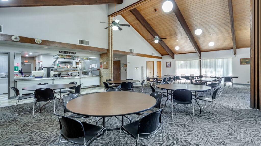 A spacious dining area with round tables and black chairs, featuring a kitchen in the background and large windows for natural light.