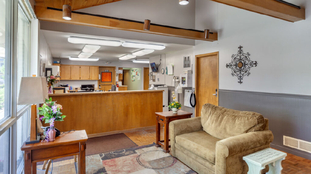 A cozy reception area featuring a brown couch, wooden reception desk, and decorative wall art. Brightly lit with large windows.