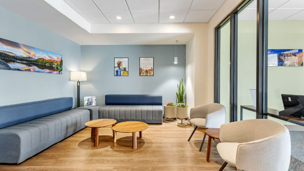 A modern waiting area featuring gray and blue sofas, wooden tables, plants, and framed artwork on the walls.