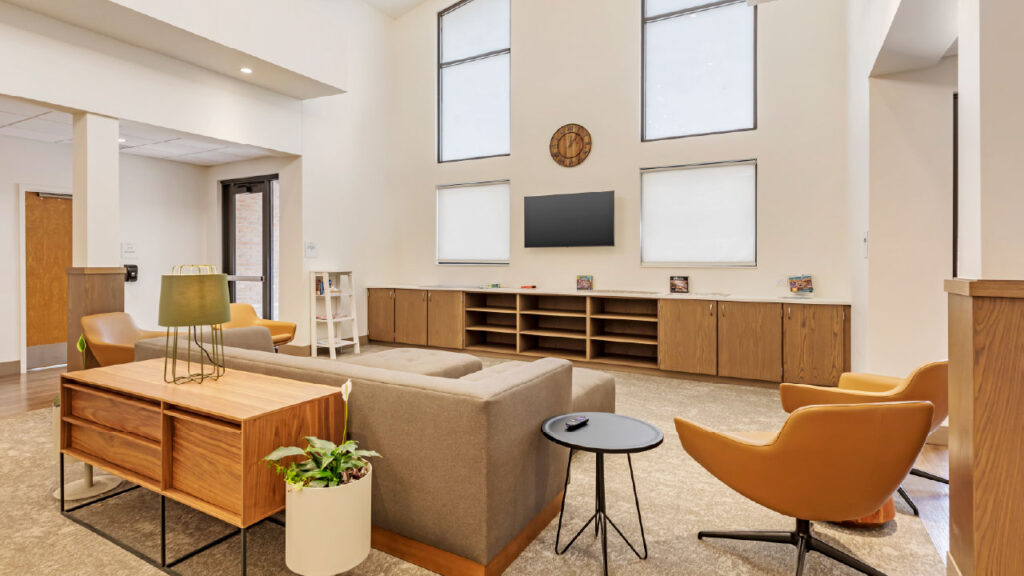 A modern living area featuring a gray sofa, orange chairs, a wooden coffee table, and a TV on the wall, with large windows for natural light.