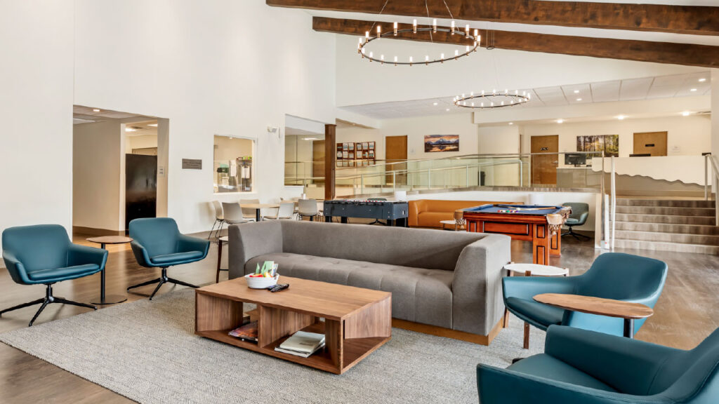 A modern lounge area featuring a gray sofa, teal chairs, a wooden coffee table, and game tables, with large windows and wooden beams.
