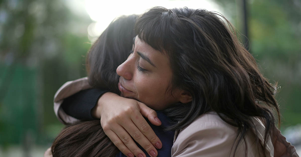Two women embrace warmly, sharing a moment of connection in a natural setting. Soft light enhances the emotional atmosphere.