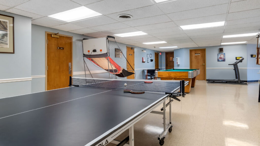 A recreational room featuring a ping pong table, pool table, treadmill, and various doors. Brightly lit with a clean floor.