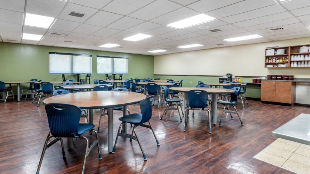 A spacious dining area with round tables and blue chairs, featuring large windows and a serving counter against a green wall.