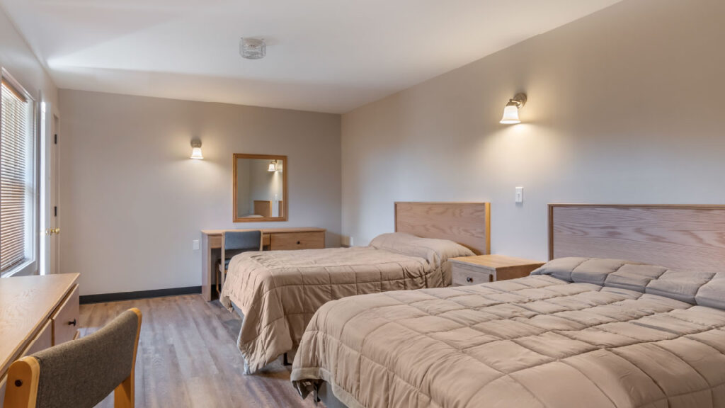 A simple bedroom featuring two beds with beige comforters, a wooden dresser, and a mirror. Natural light from a window.