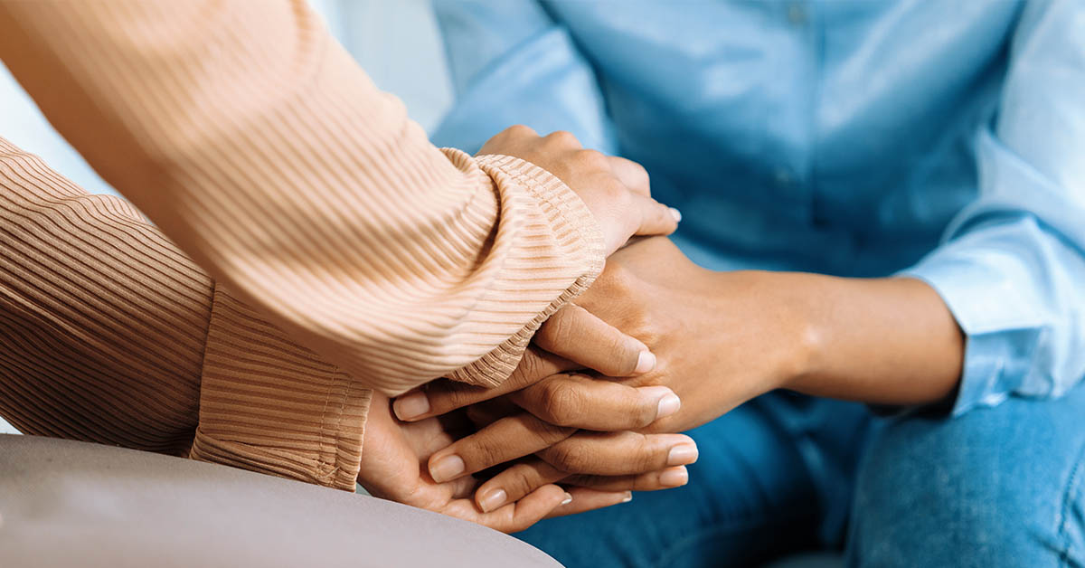 Two pairs of hands are gently clasped together, symbolizing support and connection, with one hand resting on top of the other.