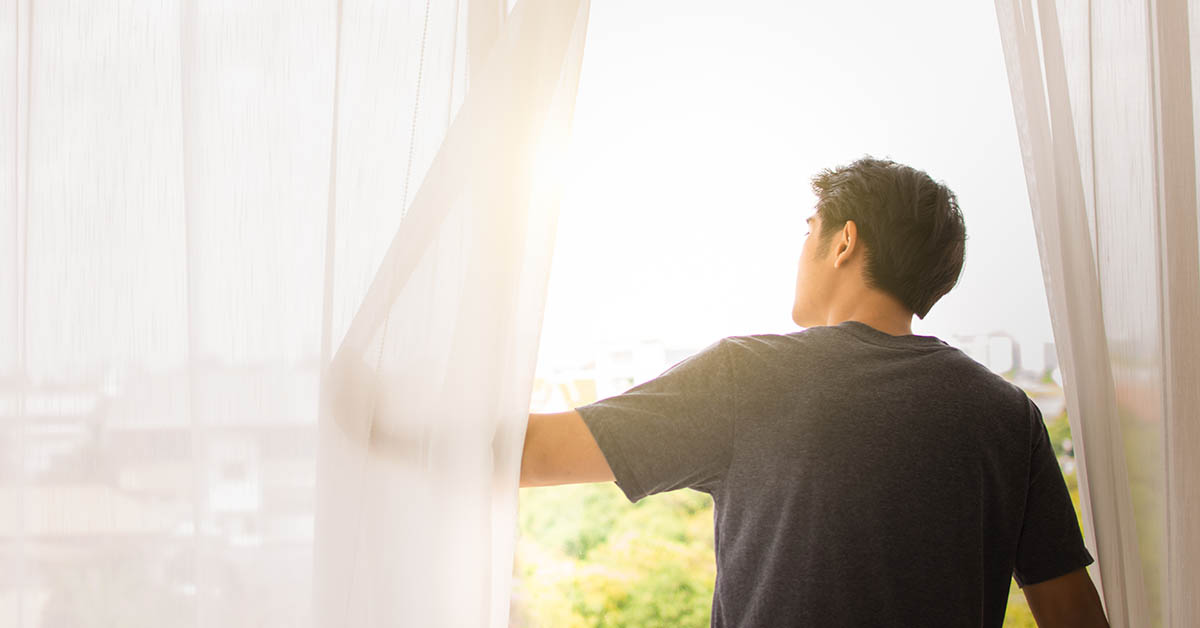 A person stands by a window, pulling back sheer curtains to let in sunlight, with greenery visible outside.