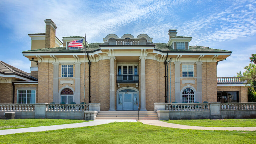 A large, elegant brick mansion with a green roof, featuring columns, a balcony, and an American flag. Lush grass surrounds the entrance.
