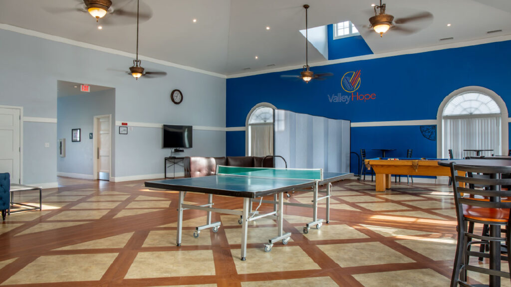 A spacious recreational room with a ping pong table, seating areas, and a pool table, featuring blue walls and natural light.