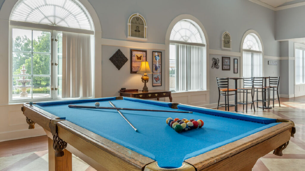 A bright room featuring a pool table with blue felt, a lamp, and a bar table. Large windows provide natural light and a view of greenery outside.