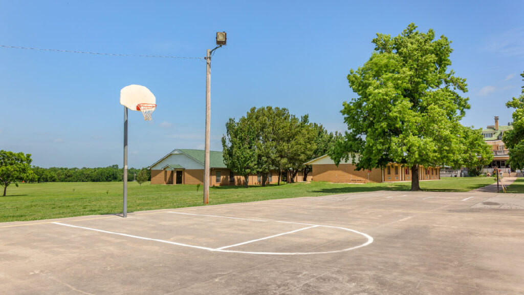 A basketball hoop stands on a court with a clear blue sky, surrounded by green grass and trees, near a building.