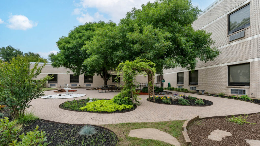 A landscaped courtyard featuring a circular path, greenery, a fountain, and seating areas surrounded by brick walls.