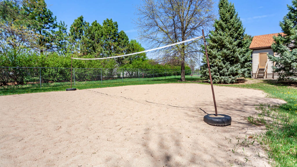 A sandy volleyball court with a net, surrounded by trees and a fence, next to a building. Clear blue sky above.