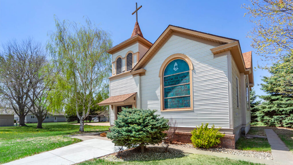A quaint church with a cross on top, surrounded by trees and greenery, featuring a decorative window with text.